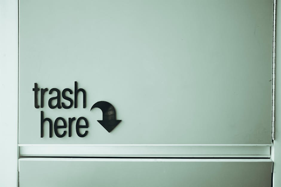 Image of an overflowing communal rubbish bin situated on a paved urban street, surrounded by various discarded waste items. The metal bin, labeled for mixed paper and cardboard recycling, has its lid