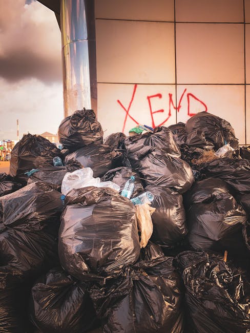 An outdoor area with a dirt and gravel surface scattered with small pieces of mixed waste materials, including plastic, paper, and textiles. Behind this, a long row of large, irregularly shaped rubbis