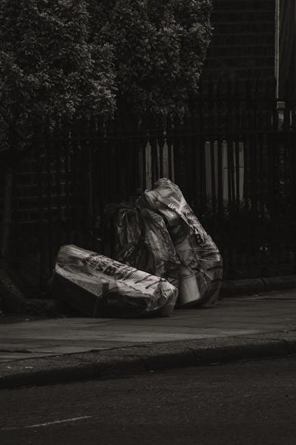 Two large black garbage bags, made of heavy plastic material with crinkled textures, are placed on a sidewalk in front of a wooden fence with vertical slats. The bags appear to be filled with househol
