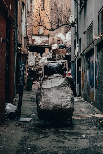 Two large black garbage bags, made of heavy plastic material with crinkled textures, are placed on a sidewalk in front of a wooden fence with vertical slats. The bags appear to be filled with househol