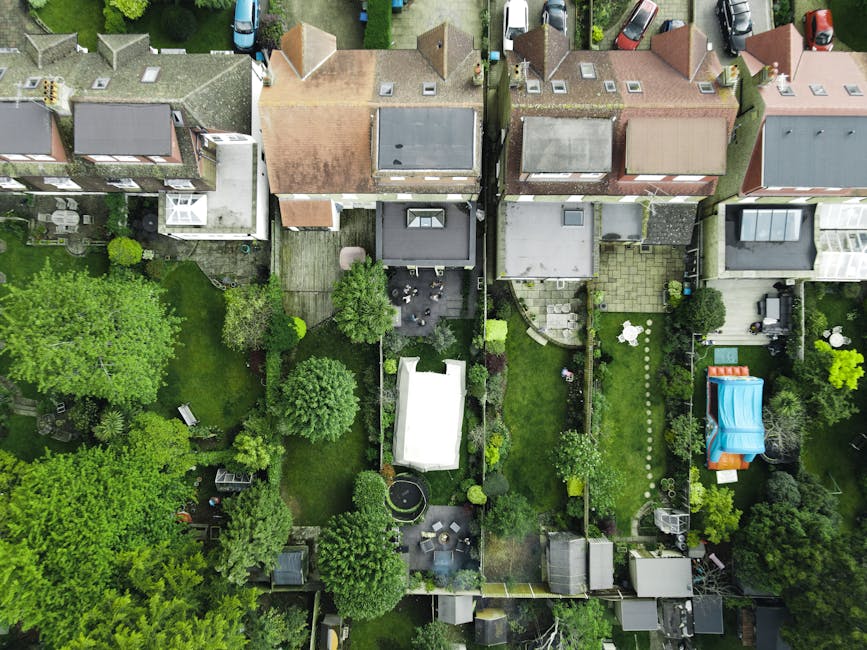 An aerial view of a row of residential houses with varied roof styles and colors, including pitched roofs with tiles and flat roofs, each with well-maintained back gardens. The gardens contain a mix o